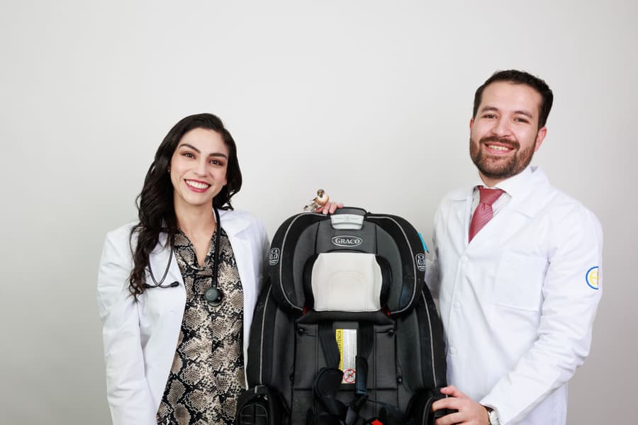 La Dra. Mariana Macías y el Dr. Ramsés Zúñiga en bata blanca junto a una silla portabebés para automóvil.
